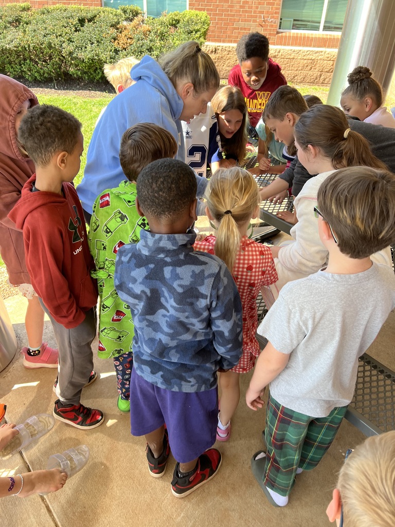 A group of elementary school children gathered around a table outdoors, closely observing something with an adult who is guiding them.