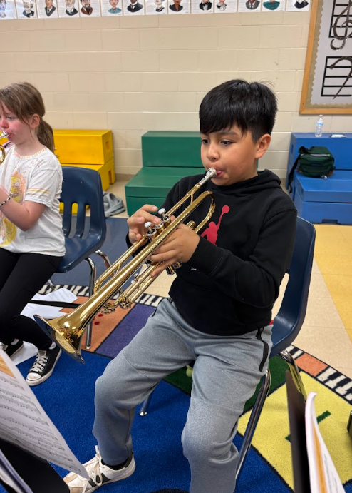A fifth grade student sits in a music classroom playing a trumpet, focused on their sheet music while seated on a chair on a colorful rug.