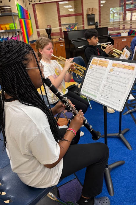 Several fifth grade students sit in a music classroom playing instruments, including clarinet and trumpet, while reading from sheet music on stands.