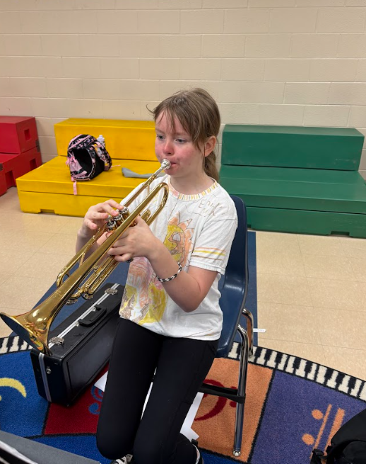 A fifth grade student sits and plays a trumpet in a classroom, concentrating on their instrument with a case and colorful seating blocks behind them.