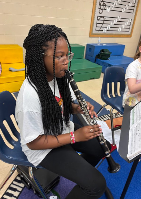 A student sits in a music classroom playing a clarinet, holding the instrument upright while reading sheet music on a stand nearby.