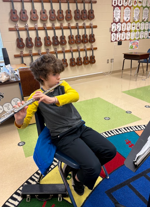 A student sits on a chair in a music room playing a flute, holding the instrument horizontally while looking toward a music stand.