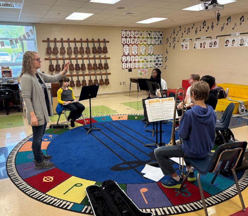 A band director stands at the front of a music classroom conducting a small group of fifth grade students seated in a circle with music stands and instruments.