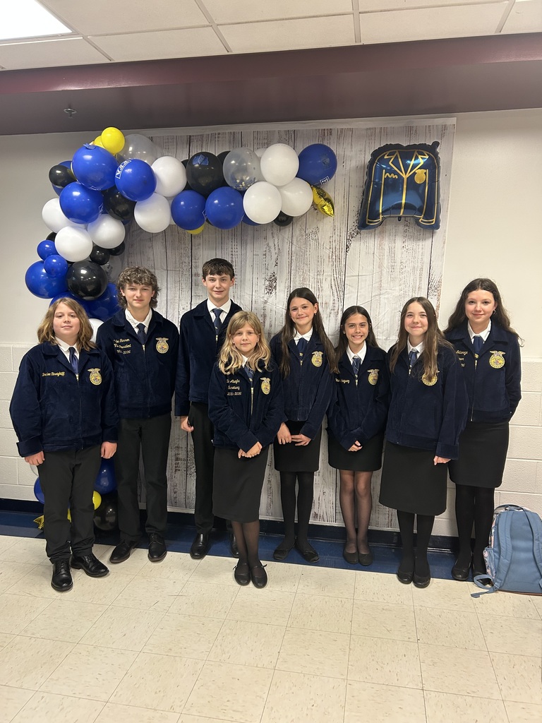 A group of eight middle school students wearing official FFA blue jackets stand side by side in front of a decorative backdrop. The backdrop features a rustic wood-style panel, a blue, white, black, and gold balloon arch, and an FFA jacket-shaped balloon. The students are dressed in formal attire—dark skirts or pants with dress shoes—and are smiling at the camera. The setting appears to be indoors, likely at a school banquet or awards event.