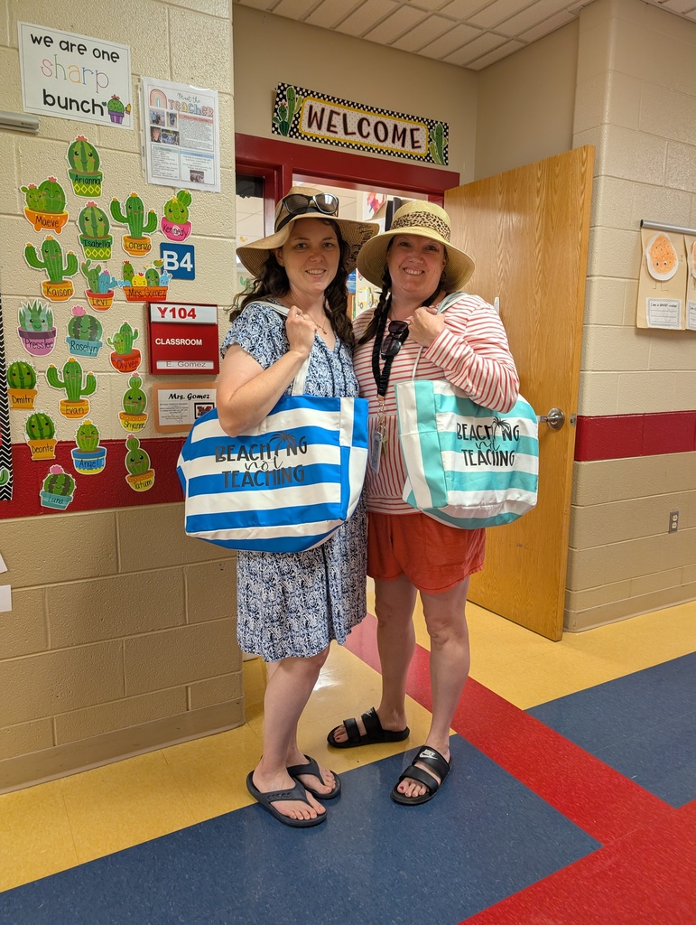 Two teachers stand in a school hallway outside a classroom doorway, smiling and dressed in beach-themed outfits. Both wear wide-brimmed sun hats and carry large striped tote bags that read “Beach Not Teaching.” One wears a blue patterned dress, and the other wears a striped top with red shorts and sandals. Classroom signs, student name displays, and a “Welcome” sign are visible on the walls.