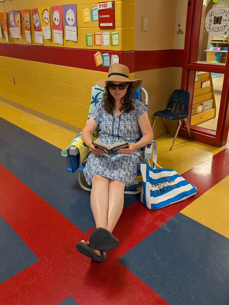 A teacher sits in a folding beach chair in a school hallway, wearing a sun hat, sunglasses, and a blue patterned dress. She has her legs crossed and is reading a book, with a striped tote bag beside her that says “Beach Not Teaching.” Classroom decorations and student work line the wall behind her.