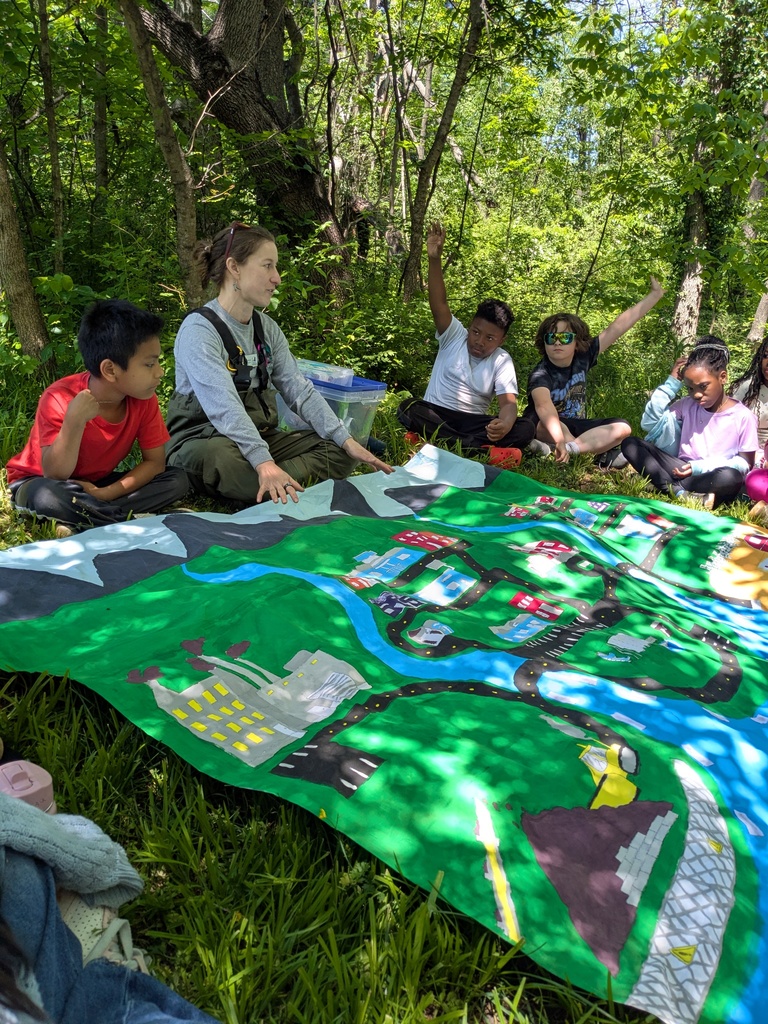 Students sit in a circle outdoors with an instructor, looking at a large illustrated map showing a watershed and surrounding community.