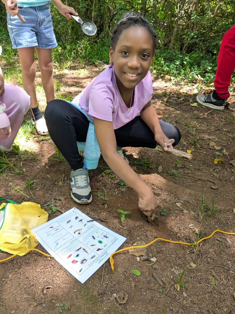 A smiling student crouches outdoors, using a stick and magnifying glass to explore soil, with an insect identification sheet nearby.