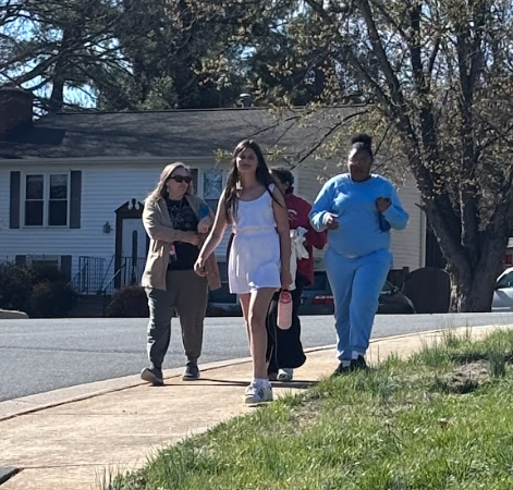 Four individuals walk on a sidewalk, with a house and trees in the background. The woman in white holds a pink bottle.