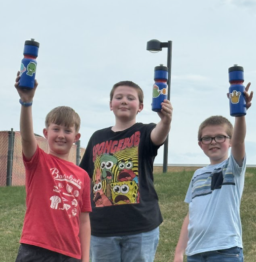Three boys in a field hold blue bottles aloft; one wears a red shirt, another a black shirt with cartoon characters, and the third a blue shirt.