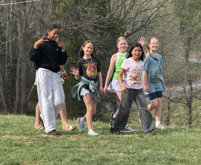Five children wearing casual clothes walking and dancing on a grassy field with trees in the background.