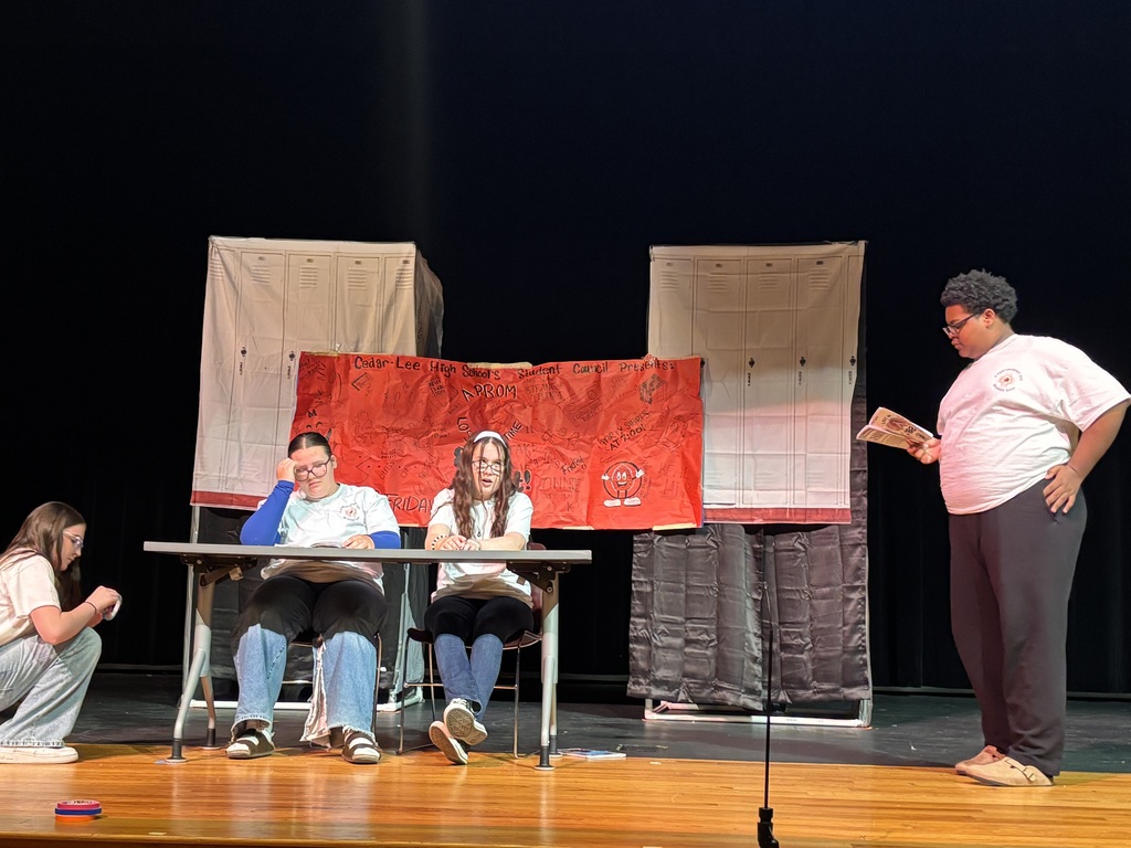 Four individuals are on stage with a microphone and table; three are seated while one stands, reading from a paper.
