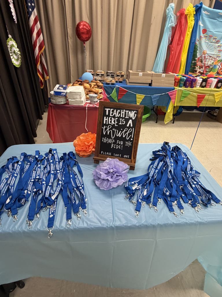 A table displays blue lanyards. Behind it, a chalkboard reads "Teaching Here Is A Miracle." Other tables have balloons, flags, and various items.