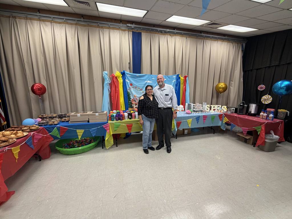 Two people stand by a table with food and balloons. The room has tables, curtains, and balloons.
