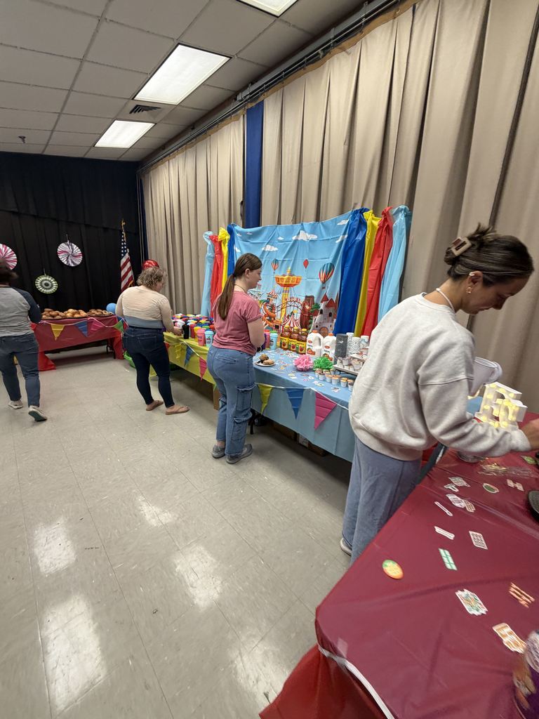 Indoor event with tables; three people preparing. Colorful tablecloths and items on tables. One person in white shirt.