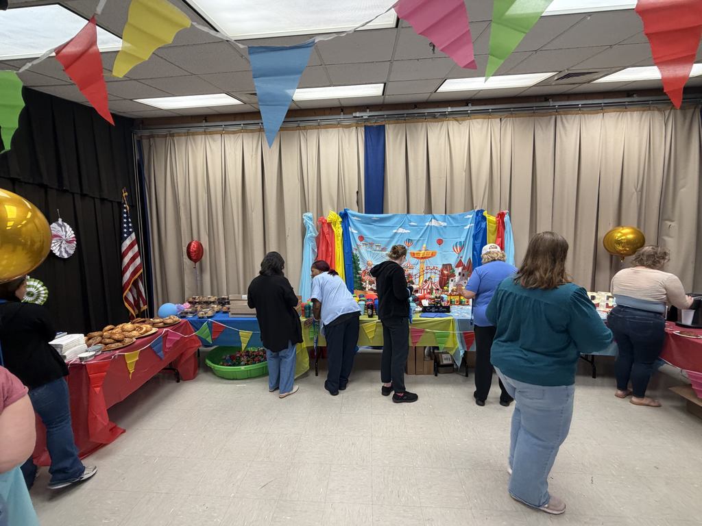 People stand around tables with food and decorations. A stage has a colorful banner. Bright balloons and flags hang from the ceiling.