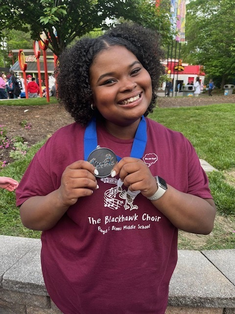 A person with curly hair holds a medal in front of a park with trees. They wear a maroon shirt.