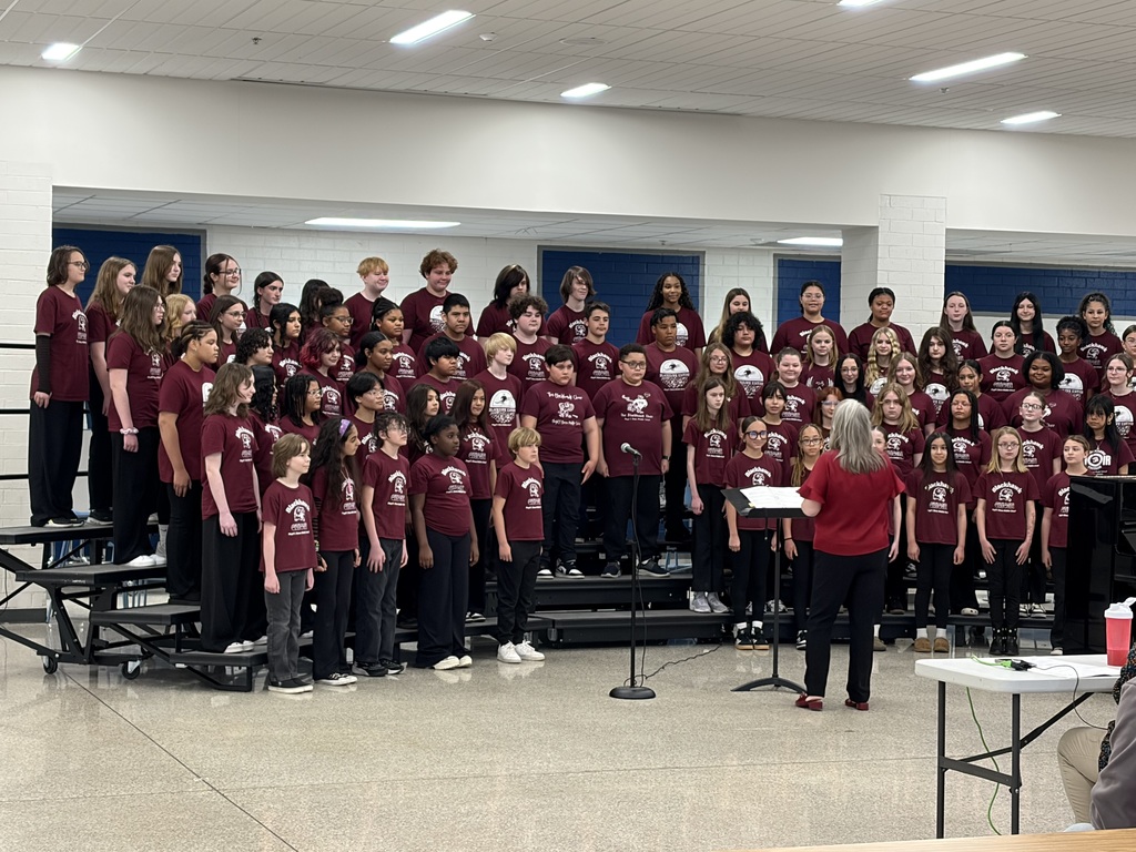 A large group of children in red shirts, likely singing, with a woman standing in front.