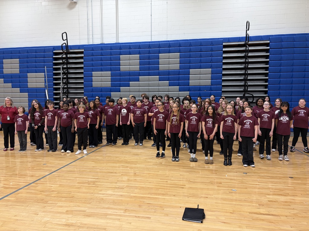Group of students in maroon shirts and black pants, standing in rows on a basketball court.