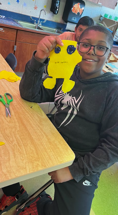 A young boy in a classroom holds a yellow paper craft of a monster, wearing glasses. Behind him, a person sits at a sink. A desk holds scissors.