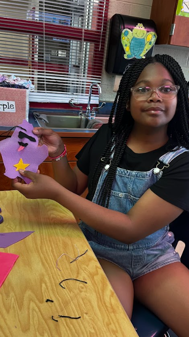 Girl with glasses, braids, and overalls holds purple paper cutout with a star. Table with papers and scissors.