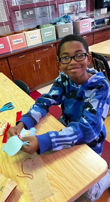 A student wearing a blue hoodie and glasses sits at a table, smiling while holding a blue paper craft.