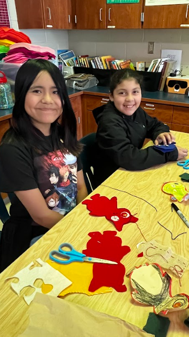 Two girls sit at a table with craft supplies, smiling. The table has scissors, paper cutouts, and other craft materials. Behind them are wooden cabinets and a shelf with books and a jar.