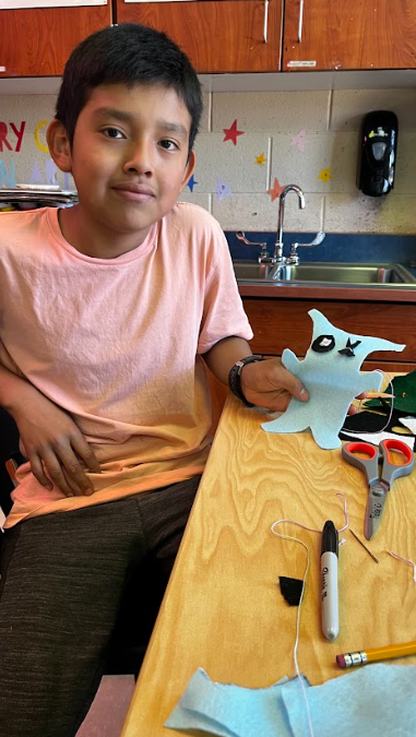 A boy in a pink shirt sits at a wooden table holding a blue craft. Items include scissors, a marker, and a pencil on the table. Behind him, a sink and cabinets are visible.
