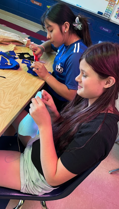Two girls seated at a table. One holds scissors, the other holds thread. Both work on crafts.