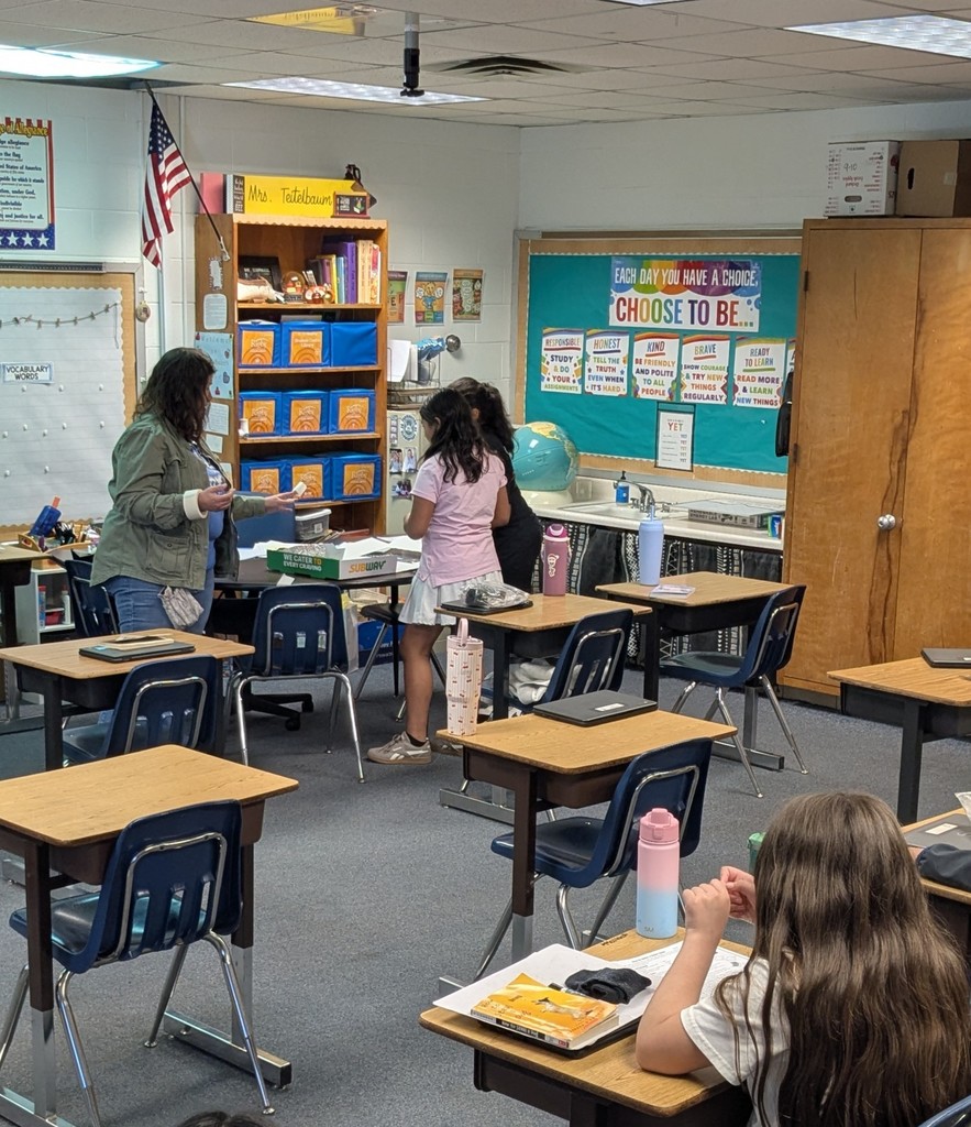 A classroom with a globe, bulletin board, and shelves. Two women converse near a whiteboard. Students sit at desks.