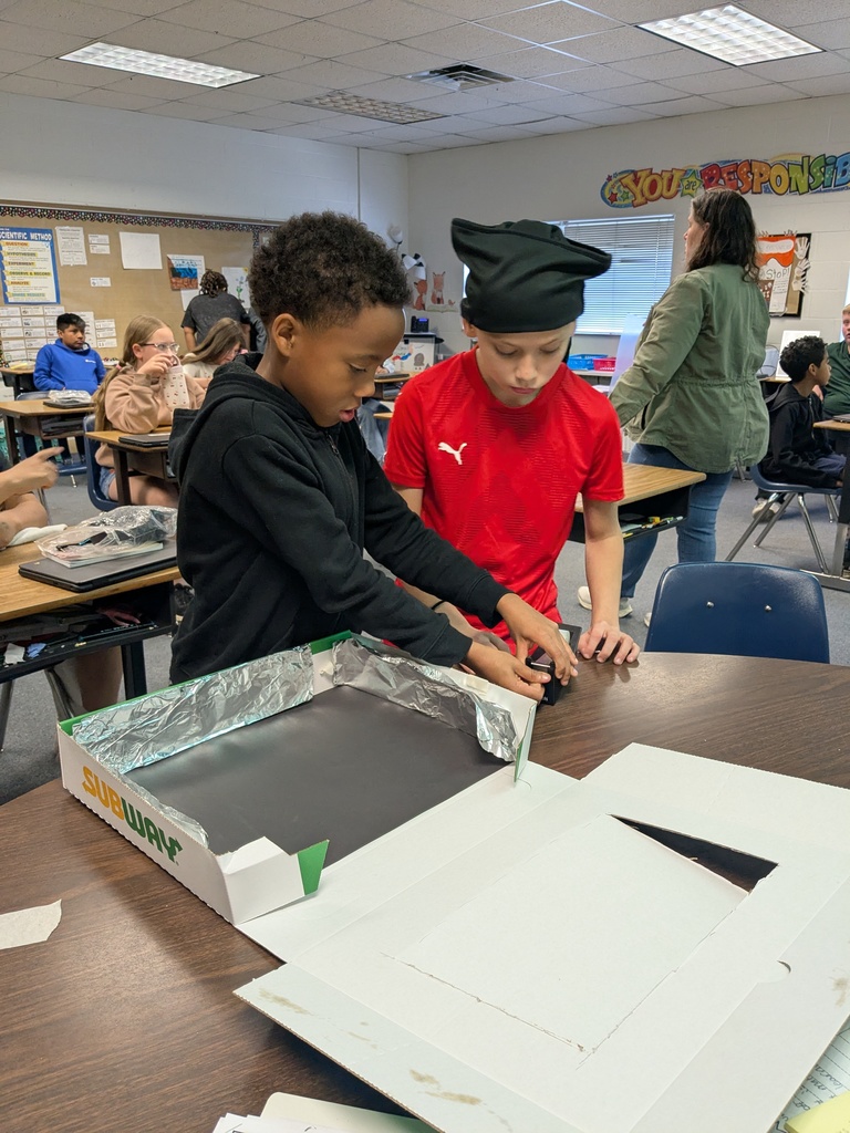 Two children in a classroom are opening a box with a metal sheet inside. Other children are seated at desks in the background.