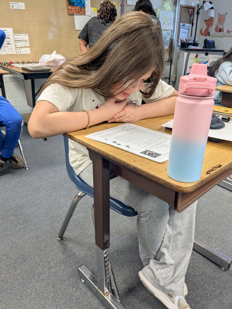 A student, wearing glasses, sits at a desk, resting her head on her arms. A pink and blue water bottle is on the desk.