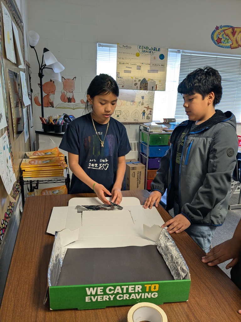 Two students open a green box on a table in a classroom. They are surrounded by school supplies.