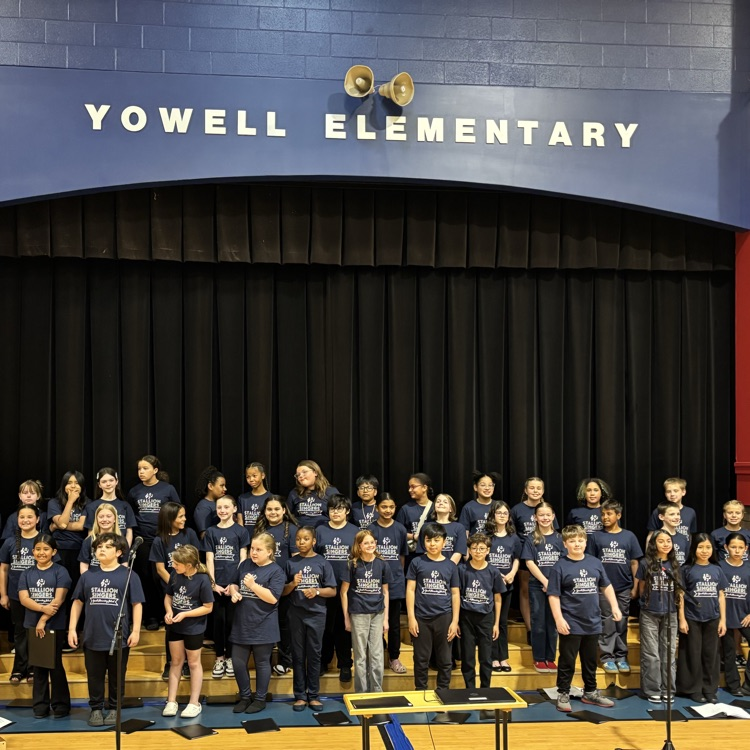 A large group of fourth and fifth grade students in blue T-shirts that read stallion singers pose on a stage after their spring concert