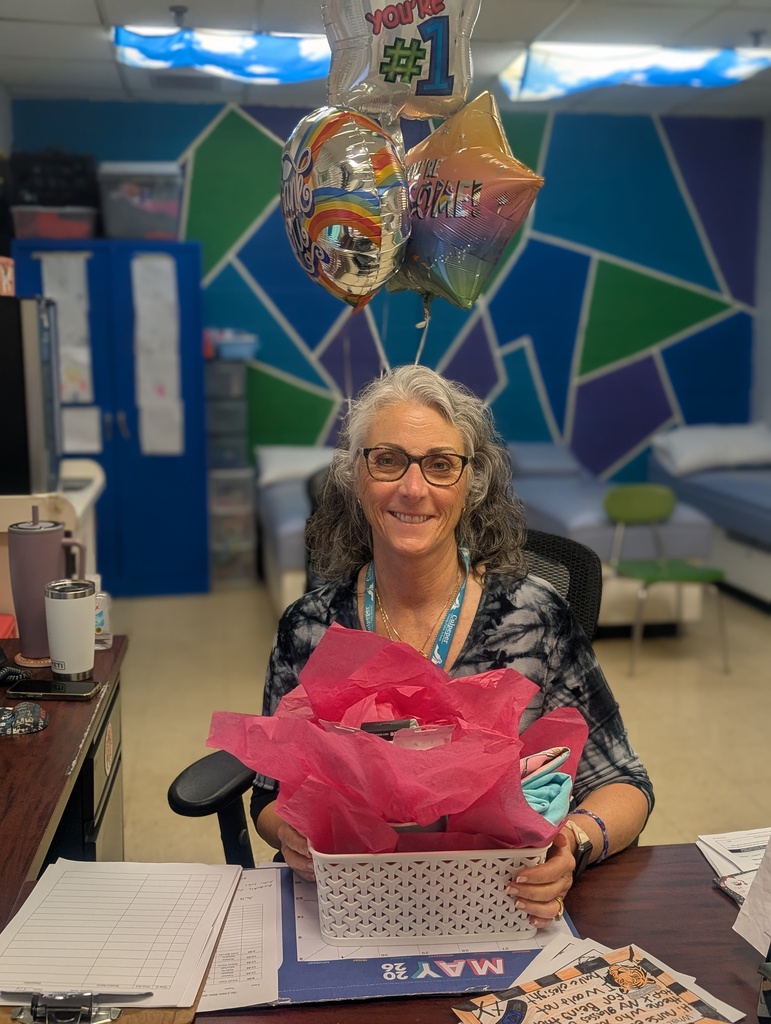 A woman with glasses sits at a desk holding a pink gift basket with a balloon bouquet.