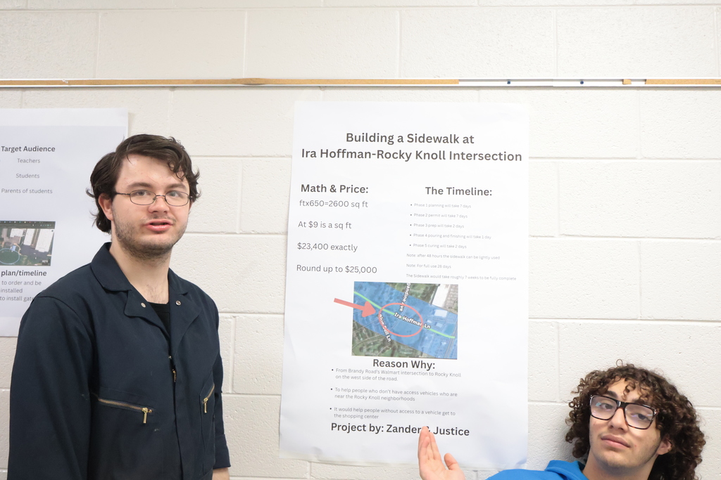 Two individuals, one in blue, one in black, pose next to a poster about sidewalk construction.
