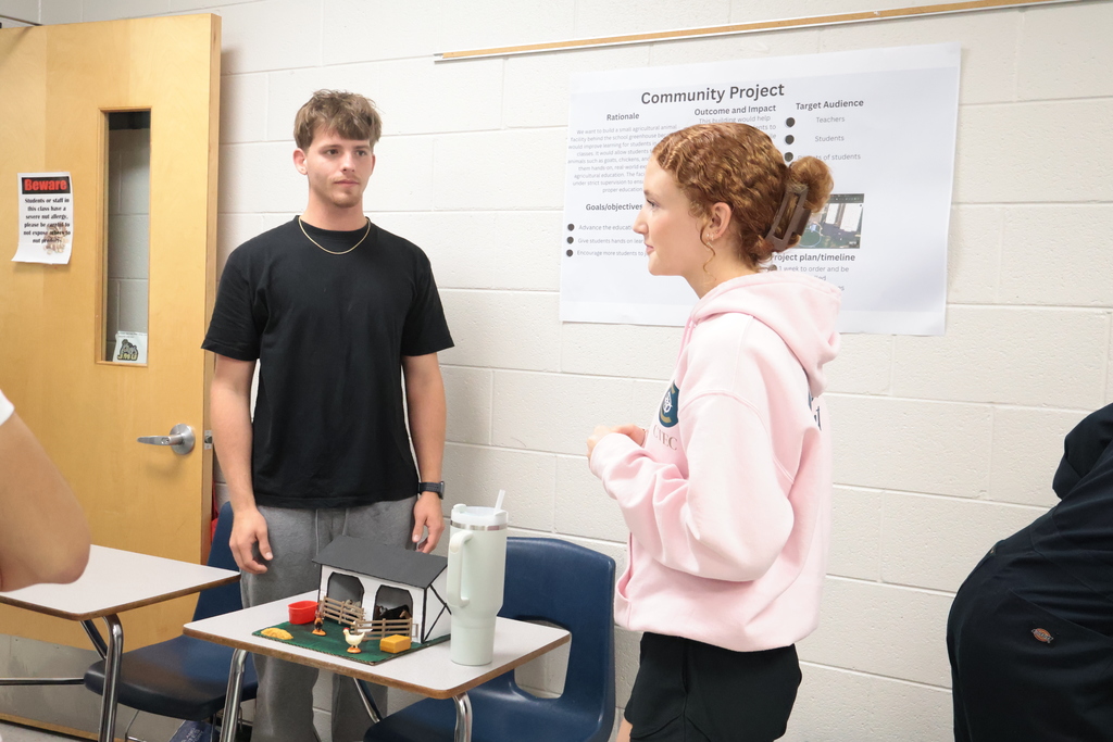 A male and female standing by a small table with a model on it. A white poster on the wall behind them.