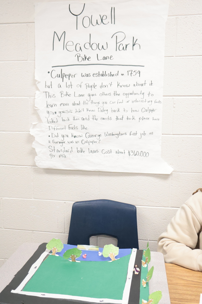 A classroom with a wall displaying a torn paper sign reading "Yowell Meadow Park Bike Lane." A table holds a small model landscape with trees.