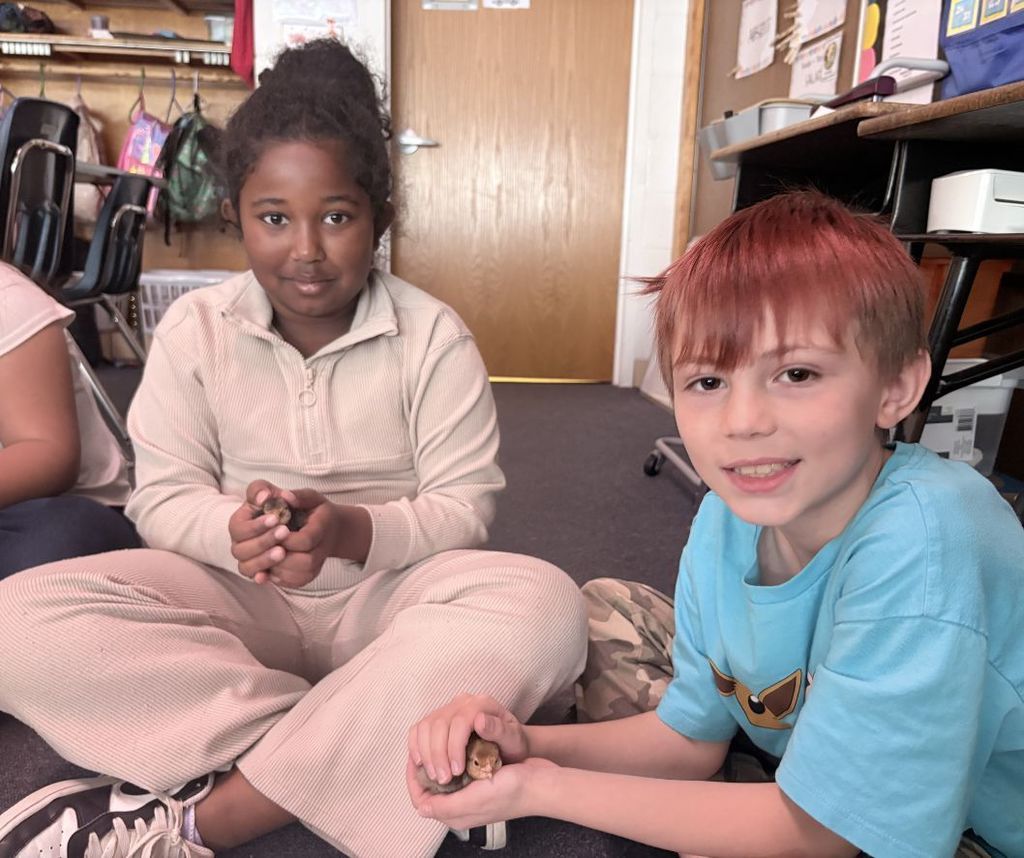 Two children sit cross-legged on the floor in a classroom, one holding a small object.