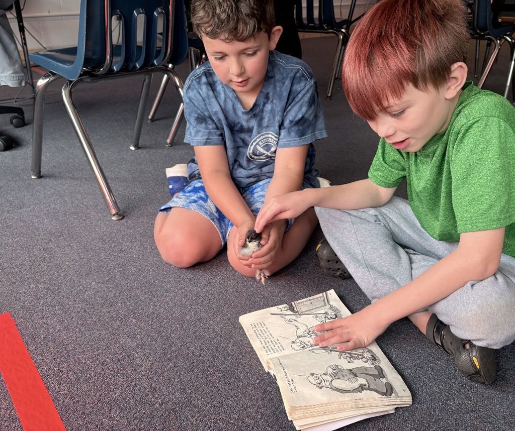 Two young boys are seated on the floor, studying an open book. One boy holds an object, and both look engaged.