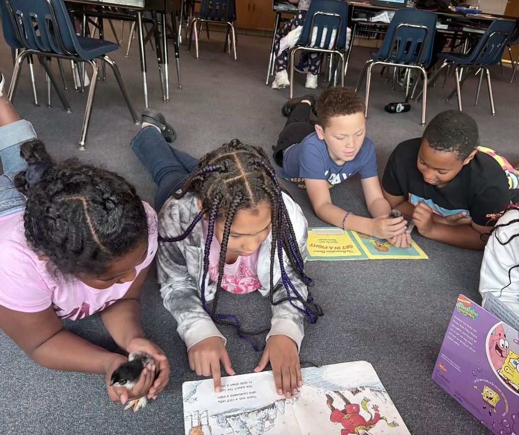 Four children reading on the floor in a classroom. One holds a book and another holds a tablet.