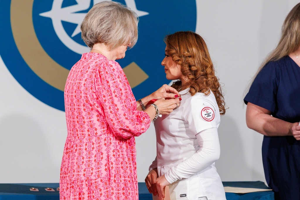 A woman in pink assists another woman in a white uniform by pinning on a badge. They stand before a white backdrop with a logo.
