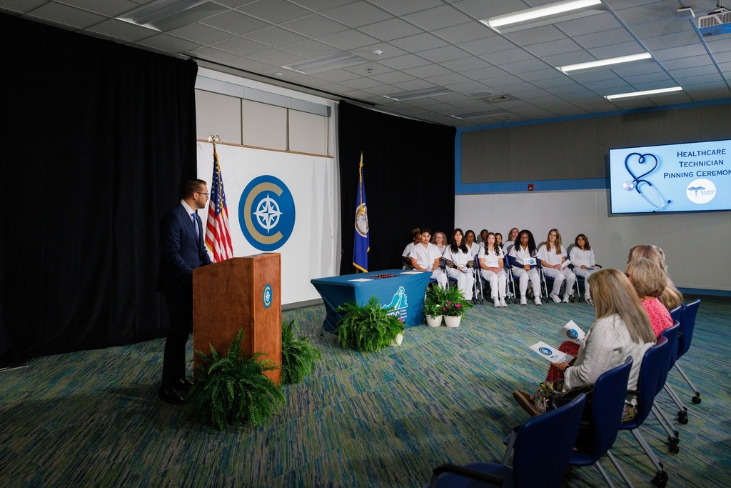 A speaker stands at a podium in front of an audience seated in chairs. Behind the speaker, a large screen displays information.