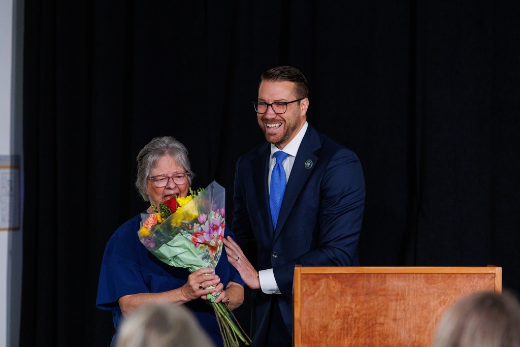 A woman with glasses holds flowers next to a man in a blue suit at a podium.
