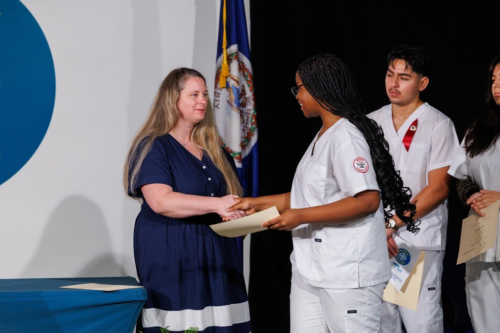 People at a ceremony. A woman in blue and a woman in white are exchanging papers. A flag is behind them.
