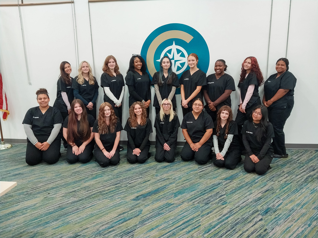 Group of CTEC students in black scrubs pose for a class photo in front of a large CTEC logo on the wall, with some students standing and others kneeling in the front row.