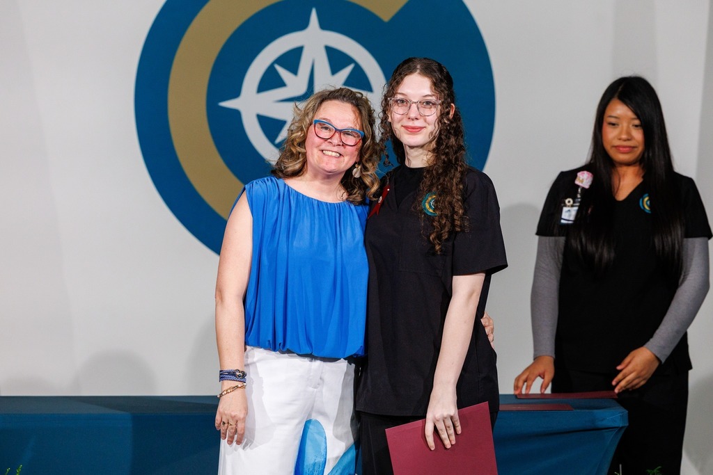 Three women stand next to each other. One holds a folder and a badge, one wears glasses.