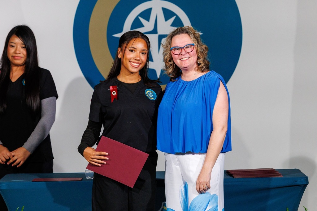 Three women stand in front of a logo; the middle woman holds a folder and a diploma.