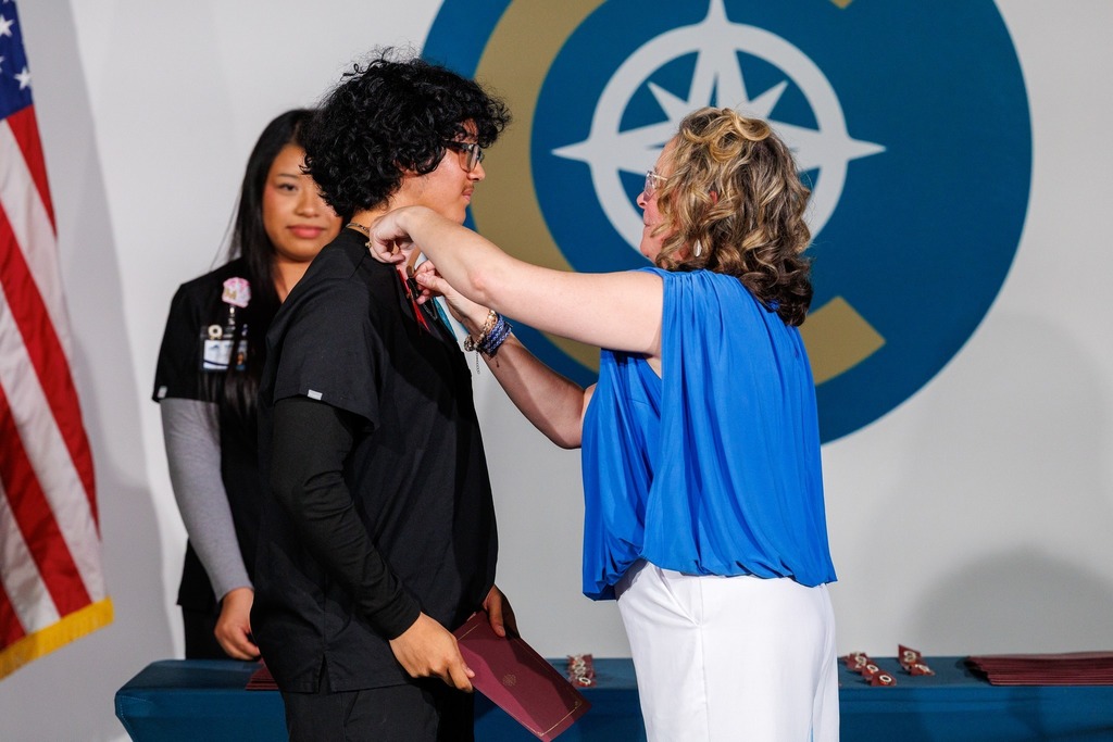 Woman pinning a badge on another woman's shoulder in a ceremony, a third woman looking on, an American flag behind.