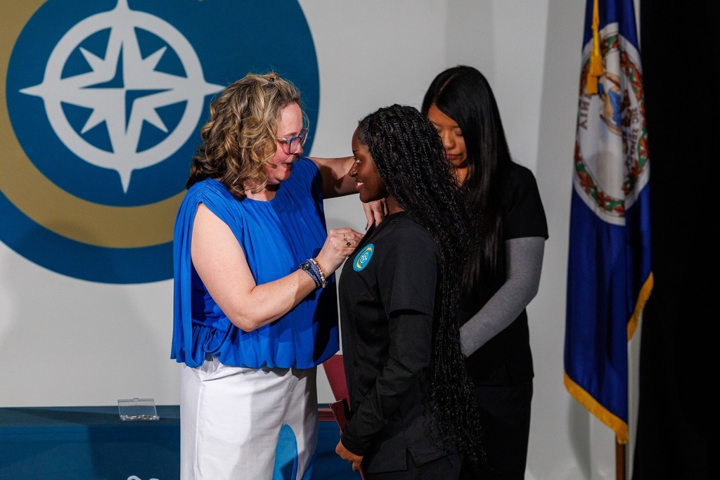 Three women stand together, with one woman placing an award on another's shoulder. They are in front of a logo and a flag.